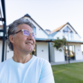 Smiling caucasian senior man wearing eyeglasses looking away against house in yard after learning about types of mortgages that would help him achieve homeownership.